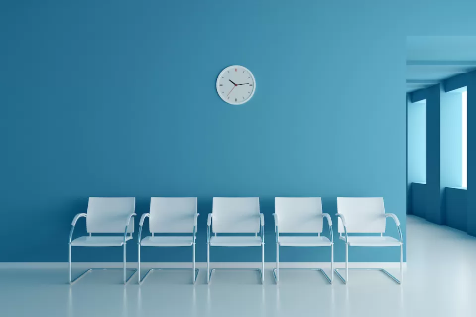 Row of five white chairs set against a blue wall with a round white analog clock. Clinic waiting room.